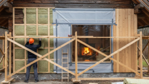 Grafika przedstawia elewację klasycznego domu z grubych, ciemnych bali w trakcie prac termomodernizacyjnych